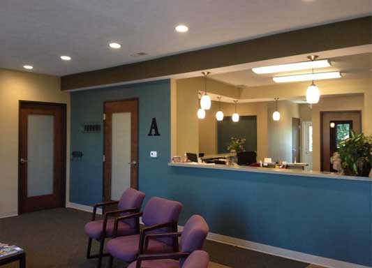 The image shows an interior space that appears to be a waiting area or reception room within a building, featuring a blue wall, a white counter with a sink and a mirror, a light-colored wooden door, chairs, and a desk with a computer monitor.