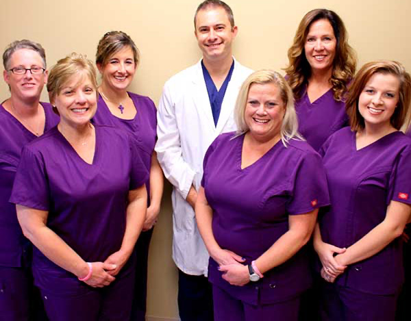 The image is a photograph of a group of people, likely a team of healthcare professionals, posing for the camera. They are dressed in scrubs and standing in front of a wall with a person seated at a desk.