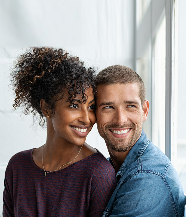 The image is a photograph of a man and woman embracing, smiling, and looking at the camera. They appear to be in an indoor setting with natural light.