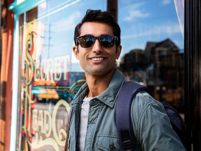 The image shows a man standing outdoors, wearing sunglasses and a backpack, with a smile on his face, posing in front of a shop window.