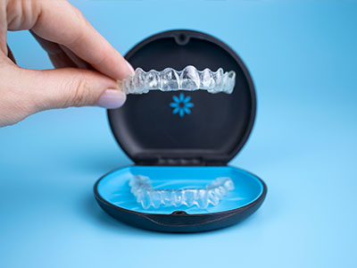 The image shows a person s hand holding an open box containing a single transparent dental retainer, with the retainer placed on top of the box to demonstrate its shape.