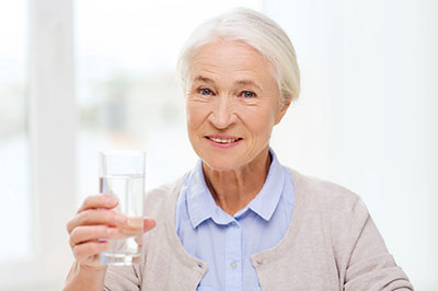 An elderly woman is holding a glass of water, smiling at the camera.