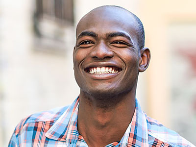 The image shows a man smiling broadly, with his eyes closed and teeth showing. He has short hair and is wearing a blue plaid shirt.