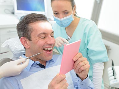 A man is holding a pink card with a surprised expression while seated in an oral surgeon s chair, surrounded by dental equipment and professionals.