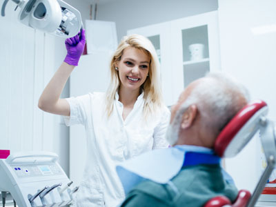 A dental hygienist, wearing a white uniform and purple gloves, is assisting an elderly man with a medical device in what appears to be a dental office.