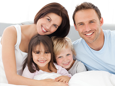 A family of four, including two adults and two children, lying on a bed with smiles, looking at the camera.