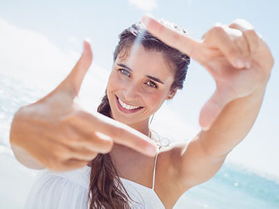 A young woman taking a selfie with her hand in front of her face, set against a sunny beach backdrop.