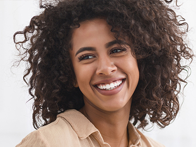 The image depicts a smiling woman with curly hair, wearing a cream-colored top and standing against a light background.